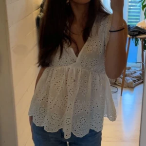 Vit broderad blus med v-ringning - Supersöt vit blus med v-ringning och broderade detaljer. Blusen är ärmlös och har en luftig, lätt utsvängd passform med fina hålbroderier och vågig nederkant. Perfekt för varma dagar och ger en romantisk vibe till din outfit.