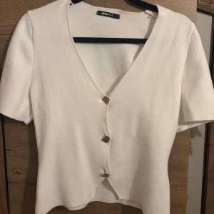Vit v-ringad topp från Gina Tricot - Vit v-ringad topp från Gina Tricot med korta ärmar och guldknappar framtill. Toppen är lätt stickad känsla och fint skick🫶🏻
