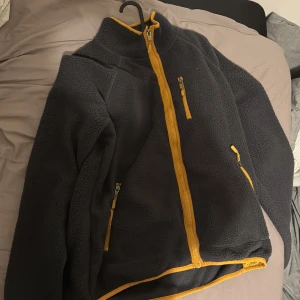 Mörkgrå fleecejacka med gula detaljer - Säljer en mörkgrå fleecejacka med snygga gula dragkedjor och kantband. Jackan har hög krage och tre praktiska fickor med dragkedja. Perfekt för kyliga dagar och enkel att matcha med din vardagsstil.