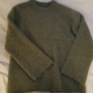 Olivgrön fluffig stickad tröja XS - Supermjuk och fluffig olivgrön stickad tröja från Stockholm Studio. Tröjan har rund halsringning och långa ärmar, perfekt för lager på lager under kyliga dagar. Den har en oversized känsla och är gjord i ett gosigt material som håller dig varm.