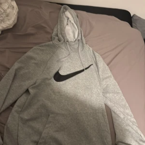 Grå Nike hoodie med svart logo - Säljer en grå hoodie från Nike med stor svart swoosh-logga på bröstet. Tröjan har huva med snörning och en klassisk känguruficka framtill. Perfekt för chill dagar eller när du vill ha en sportig look.