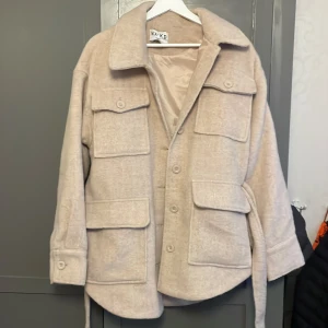 Beige mellanlång jacka från NA-KD - Säljer en beige jacka från NA-KD i storlek 38. Jackan har fyra stora fickor framtill, knappar hela vägen och ett bälte i midjan. Snygg krage och långa ärmar med knapp vid mudden.