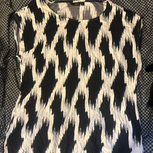 Svartvit mönstrad klänning från Lindex - Cool svartvit topp från Lindex med ett unikt, abstrakt mönster. Modellen är ärmlös och har en rak, avslappnad passform. Perfekt för dig som gillar att sticka ut med grafiska prints. Tillverkad i mjuk bomull som känns skön mot huden.