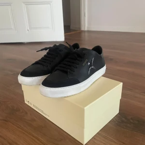 Svarta Axel Arigato sneakers - Snygga svarta sneakers från Axel Arigato med vit sula och svart snörning. Skorna har en stilren design med fågel-logga i silver på sidan och detaljer i guld vid snörningen. Perfekta för dig som gillar minimalistisk och trendig stil. Använda 2 gånger.