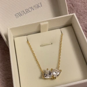 Swarovski halsband guld - Snyggt halsband från kollektionen ”mesmera” från Swarovski. Två gnistrande hjärtformade vita stenar på en tunn kedja med guldfärgad plätering. Perfekt för dig som gillar eleganta detaljer och vill ha något som sticker ut. Aldrig använt och kommer i orginal ask.
