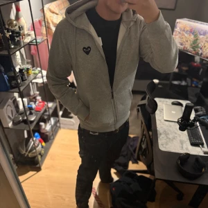 Zip Hoodie Comme des Garçons Play - Grå hoodie med dragkedja från Comme des Garçons Play. Har huva med snörning, känguruficka och svart hjärta med ögon på bröstet och ryggen. Perfekt för dig som gillar streetstyle och vill sticka ut med en ikonisk detalj.