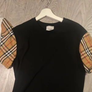 Burberry t-shirt  - Svart t-shirt från Burberry med klassiska rutiga korta ärmar i beige, rött och svart. Snygg och stilren design med rund halsringning. Perfekt för dig som vill ha en ikonisk look med en twist. Gjord i mjuk bomull för skön känsla.