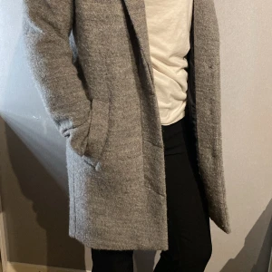 Grå ullkappa med knappar - Snygg grå kappa i ullmix med klassisk krage och tre svarta knappar framtill. Jackan har två fickor på sidorna och är helfodrad med svart tyg. Perfekt för dig som gillar stilrena looks. 