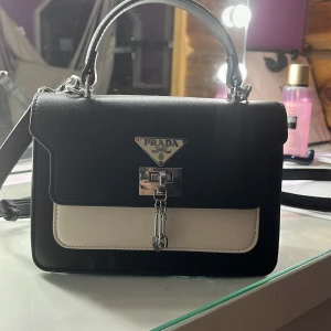 Svart och vit handväska från Prada - Snygg svart och vit handväska från Prada med klassisk triangel-logga framtill. Väskan har både handtag och axelrem, samt ett elegant lås med kedjedetalj. Tillverkad i skinn och perfekt för digsom vill ha en stilren accessoar med lyxig känsla.