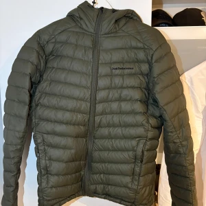 Grön dunjacka Peak Performance - Säljer en mörkgrön dunjacka från Peak Performance med huva och dragkedja framtill. Jackan har quiltad design och är perfekt för kyliga dagar. Snygg, sportig look med diskret logga på bröstet och två sidofickor med dragkedja.