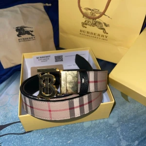 Burberry rutigt skärp med B-spänne - Snyggt Burberry-bälte i klassiskt beige, svart och rött rutigt mönster. Skärpet har ett stort metallspänne med Burberrys ikoniska B-logga i guld eller silver. Tillverkat i skinn med svart insida och exklusiv känsla. Perfekt accessoar för att lyfta din outfit.105,110,115 storlek.