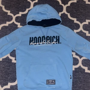 Blå hoodie från Hoodrich med tryck - Snygg ljusblå hoodie från Hoodrich med stort svart och vitt tryck på bröstet. Tröjan har huva med snörning, känguruficka och ribbade muddar. Perfekt för dig som gillar streetwear och vill sticka ut med en cool look.