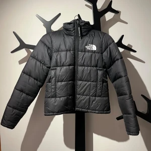 Svart pufferjacka från The North Face - Svart pufferjacka från The North Face i storlek XS. Jackan har hög krage, vita logotryck fram och bak, quiltad design och dragkedja framtill. Jackan har även en lura som går att fälla ut från kragen med hjälp av en dragkedja. Jätte bra skick.