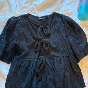 Svart topp från Gina Tricot - Supersöt svart topp från Gina Tricot med puffiga ärmar och snygg struktur i tyget. Toppen har tre knytningar framtill och ett lätt vågigt mönster som ger en cool look. Perfekt för dig som gillar unika detaljer och vill sticka ut.
