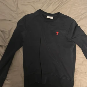 Ami äkta tröja - Svart sweatshirt från Ami Alexandre Mattiussi med broderat rött hjärta på bröstet och diskret Ami-logga i nacken. Tröjan har rund hals, lång ärm och är gjord i mjuk bomull. Perfekt för dig som gillar stilrena detaljer och fransk design.