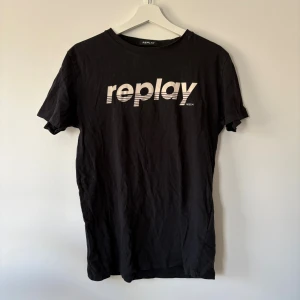 Svart Replay t-shirt med tryck - Svart t-shirt från Replay med vit logotyp i coolt grafiskt tryck på bröstet och gul patch på ärmen. Klassisk rund hals och korta ärmar. Tillverkad i mjuk bomull som känns skön mot huden. Perfekt för dig som gillar streetwear och enkel stil.