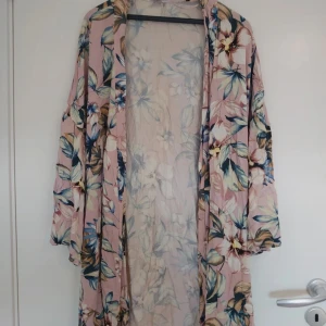 Blommig morgonrock, Secret Possessions, stl M/L - Fin morgonrock från Secret Possessions i storlek M/L. Klänningen är ljusrosa med stort blommigt mönster i blått, grönt och beige. Den har omlottmodell med knytband i midjan och långa ärmar. Perfekt att svepa om sig för en elegant look 