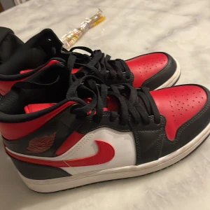Nike Air Jordan 1 Mid röd/svart/vit - Säljer ett par Nike Air Jordan 1 Mid i klassisk röd, svart och vit färg. Skorna har svart snörning, röd swoosh och detaljer, samt vit mellansula. Tillverkade i skinn med hög siluett och ikonisk Jordan-logga på sidan och plösen. Perfekta för dig som vill sticka ut med en tidlös sneaker.