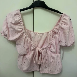 Ljusrosa blus med knyt och volang - Supersöt ljusrosa blus med broderade hålmönster och puffiga korta ärmar. Toppen har en v-ringad skärning med knyt framtill och volangdetaljer som ger en romantisk vibe. Perfekt för dig som gillar pasteller och detaljer.