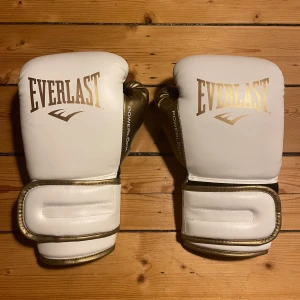 Everlast boxningshandskar - Säljer ett par snygga 10 Oz Everlast powerlock 2R i  vitt, svart och guld, har endast använt handskarna ca 3 ggr och säljer dem för dem helt enkelt är för små. Handskarna har bred kardborre och coola detaljer i guld vid tummarna och texten. Perfekta för säck och mittsar, funkar också för sparring om du är liten. Köpte handskarna för 799kr.