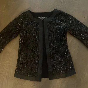 Svart paljettjacka från Zara Collection - Säljer en svart cardigan jacka från Zara Collection med glittriga paljetter över hela ytan. Jackan har rundad hals, trekvartsärm och öppen front med satinliknande kant. Perfekt för dig som vill sticka ut med lite extra bling. Passar även xs/s se bilderna🙂