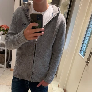 Grå kofta från Superdry med huva - Grå stickad hoodie från Superdry Premium Black Label med broderad logga på bröstet och praktisk huva. Tröjan har dragkedja framtill, ribbade muddar och två fickor. Perfekt för chill dagar och snygg till jeans. Perfa för höst och vinter