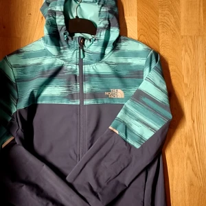 Blå och turkos vindjacka från The North Face - Snygg vindjacka från The North Face i mörkblått och turkos med coolt mönster upptill och på ärmarna. Jackan har huva med snörning, hel dragkedja och logga på bröstet. Perfekt för dig som gillar streetstyle och outdoor-vibe. Så gott som ny. använd ett par ggr men väldigt få ggr köpt för 2300. Säljer för 1200 kb