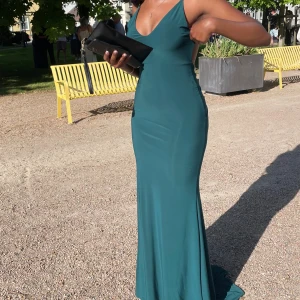 Mörkgrön långklänning med öppen rygg - Snygg mörkgrön långklänning med smala axelband och djup v-ringning. Klänningen har en öppen rygg med twistad detalj baktill. Materialet är mjukt och följsamt, perfekt för att sticka ut på bal eller fest. Endast använd en gång och är i storlek S