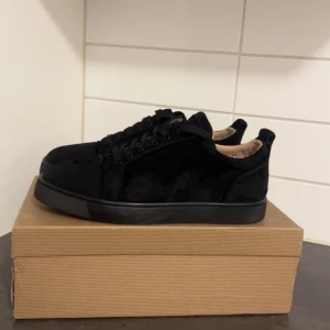 Svarta sneakers från Christian Louboutin - Snygga svarta sneakers från Christian Louboutin i mocka med klassisk röd sula och logotyp. Skorna har rund tå, platt sula och snörning. Insidan är beige och de levereras med originalkartong och dustbag. Perfekt för dig som gillar exklusiva detaljer och stilren design.