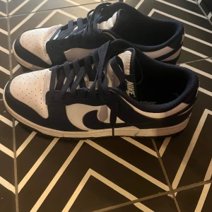 Nike Dunk Low vit/marinblå sneakers - Nike Dunk Low sneakers i vit och marinblå färg med klassisk Swoosh-logga på sidorna. Skorna har snörning, perforerad tåbox och platt sula. Ovandelen är i skinn och syntet, med lågt skaft och bekväm passform. Perfekta för dig som gillar streetwear och sportig stil.