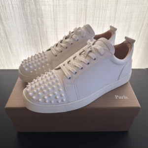 Christian Louboutin vita sneakers - Säljer ett par sjukt snygga vita sneakers från Christian Louboutin med ikoniska röda sulor med spikar. Skorna är i slätt skinn, har snörning och en rund tå. Perfekta för dig som vill sticka ut med exklusiv vibe och edgy detaljer.