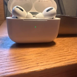 AirPods Pro - Apple AirPods Pro med trådlöst laddningsetui. Aktiv brusreducering, transparensläge och bekväm passform. Fodralet har synliga repor och mindre slitage, men fungerar som det ska. Perfekt för musik, samtal och träning.