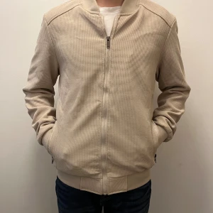 Beige bomberjacka med dragkedja - Snygg beige bomberjacka med ribbade muddar och krage. Jackan har ett diskret rutmönster i materialet och stängs med dragkedja framtill. Perfekt för dig som gillar stilren och enkel design. Långärmad och med fickor på sidorna.