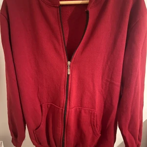 Röd hoodieLux's Brand - Röd hoodie med dragkedja från Lux's Brand. Jackan har två fickor framtill, ribbade muddar och en skön huva. Materialet är mjukt och känns som bomull, perfekt för chill dagar. Enkel och stilren design utan mönster.