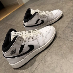 Nike Air Jordan 1 Mid   - Säljer ett par Nike Air Jordan 1 Mid sneakers i vit och svart. Klassisk design med snörning, perforerad tå och svart Swoosh på sidan. Skorna har en rund tå och är tillverkade i skinn med svart foder och detaljer. Perfekt för dig som gillar streetstyle och ikoniska sneakers. Hör av er vid eventuella frågor!!