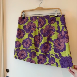 Blommig kortkjol från Zara - Superfin kortkjol från Zara med stort blommigt mönster i lila och rosa på en limegrön botten. Kjolen har en rak passform och är tillverkad i ett stickat material som känns mjukt och bekvämt. Perfekt för dig som vill sticka ut med färg och mönster.