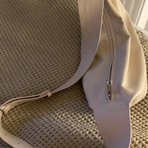 Beige axelväska  med dragkedja - Snygg beige axelväska med bred justerbar rem och silverfärgad dragkedja. . Köpt den för ganska längesen men den har mest hängt hemma och den är inte så använd. Vit lite mjölkvit eller hur man förklarar lite beige vit. Den har små skavanker som jag försökt tagit så bra bilder som möjligt på men inga stora saker bara små ytliga!