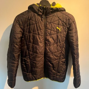 Svart quiltad pufferjacka från Puma - Svart quiltad pufferjacka från Puma med gul logga och detaljer. Jackan har huva med gul insida, dragkedja framtill och lång ärm. Perfekt för kyliga dagar och har en sportig vibe med snygg passform.