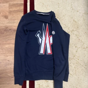 Mörkblå sweatshirt från Moncler - Säljer en mörkblå sweatshirt från Moncler med stort broderat logomärke i vitt, rött och blått framtill. Tröjan har rund hals, långa ärmar och ribbade muddar. Klassisk och stilren design med Moncler-logga även på ärmen.