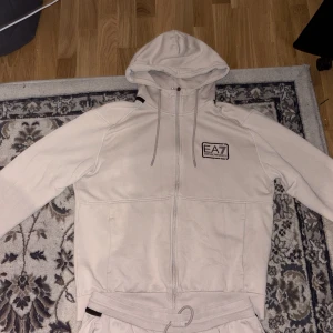 Beige hoodie från EA7 Emporio Armani - Säljer en stilren beige hoodie från EA7 Emporio Armani med dragkedja och huva. Tröjan har EA7-logga på bröstet, snörning i huvan och är tillverkad i mjuk bomullsmix. Perfekt för en chill och sportig look. Både byxorna och hoodien är storlek L, men passar även M. Pris går att diskuteras👍🏽