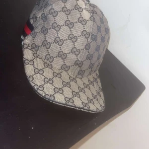 Gucci keps med GG-mönster och röd rand - Snygg Gucci keps i beige med klassiskt GG-mönster över hela kepsen. Har en röd och marinblå rand på sidan och justerbar rem baktill i svart skinn. Materialet är bomull och kepsen är tillverkad i Italien. Perfekt för dig som vill ha en lyxig streetstyle.