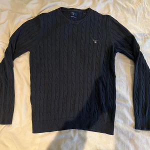 Marinblå kabelstickad tröja GANT - Snygg marinblå kabelstickad tröja från GANT i bomull. Klassisk rund hals och ribbade muddar vid ärmslut och nederkant. GANT-logga broderad på bröstet. Perfekt för dig som gillar stilrena och tidlösa plagg.