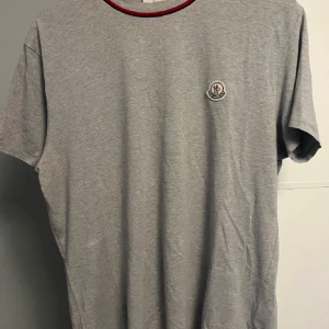 Grå Moncler t-shirt med röd kant - Snygg grå t-shirt från Moncler med klassisk logga broderad på bröstet. Rund hals med röd och mörkblå kant ger en clean och modern vibe. Tillverkad i mjuk bomull som känns skön mot huden. Perfekt för dig som gillar stilrena plagg med exklusiv touch.