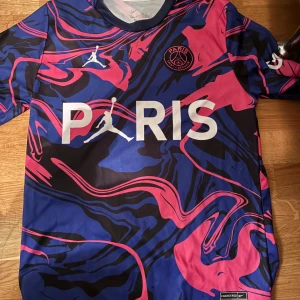 PSG Mbappé kortärmad fotbollströja - Säljer en fet PSG fotbollströja med Mbappé #7 på ryggen. Tröjan är kortärmad och har ett galet mönster i blått, svart och rosa med Paris-logga och Jordan-märke på bröstet. Tillverkad i lätt Dri-FIT material som andas, perfekt för match eller träning.