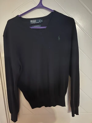 Svart v-ringad tröja Polo Ralph Lauren - Svart v-ringad tröja från Polo Ralph Lauren i 100% merinoull. Klassisk design med broderad logga på bröstet och ribbade muddar vid ärmslut och nederkant. Perfekt för dig som gillar stilrena och tidlösa plagg.