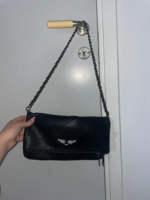 Svart axelväska med kedja - Snygg svart axelväska i skinnimitation med kedja som axelrem. Väskan har en stilren rektangulär form, dragkedja upptill och ett litet fack med dragkedja på baksidan. Den är aldrig använd så den är i helt nyskick.