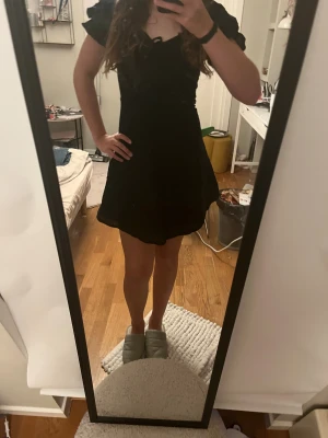 Svart kort klänning med puffärm - Supersöt svart kort klänning med puffiga kortärmar och snörning vid bysten. Klänningen har en lätt utställd kjol och är sydd i ett tunt, mjukt tyg som känns luftigt. Perfekt för dig som gillar en feminin och trendig stil.