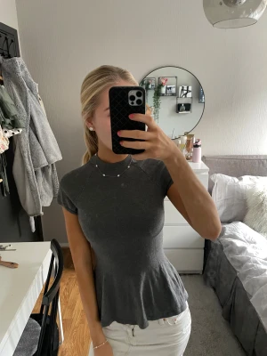 Mörkgrå ribbad topp med kort ärm - Säljer en mörkgrå ribbad topp med korta ärmar och hög hals. Toppen har en figurnära passform upptill och släpper ut i en lätt volang nertill. Materialet är stretchigt och mjukt, perfekt för en stilren look.