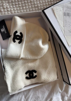 Vit mössa och halsduk från Chanel - Exklusiv vit stickad halsduk från Chanel med klassisk svart Chanel-logga broderad på ena änden. Halsduken är mjuk och har en stilren, enkel design som passar perfekt till kalla dagar. Kommer i originalbox och är en riktig statement-accessoar. Finns kvitto!!