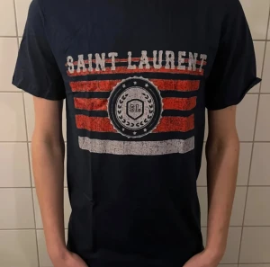 Saint Laurent t-shirt med tryck - Snygg mörkblå t-shirt från Saint Laurent med tryck i rött och vitt framtill. T-shirten har korta ärmar och klassisk passform. Materialet känns mjukt och skönt, perfekt för en avslappnad stil.